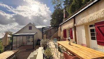 A patio with a wooden table and chairs, a greenhouse, and a building with red shutters.