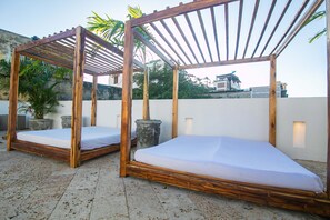 Dos estructuras de cama de madera con colchones blancos, rodeadas de plantas y una pared blanca, en una terraza en la azotea.