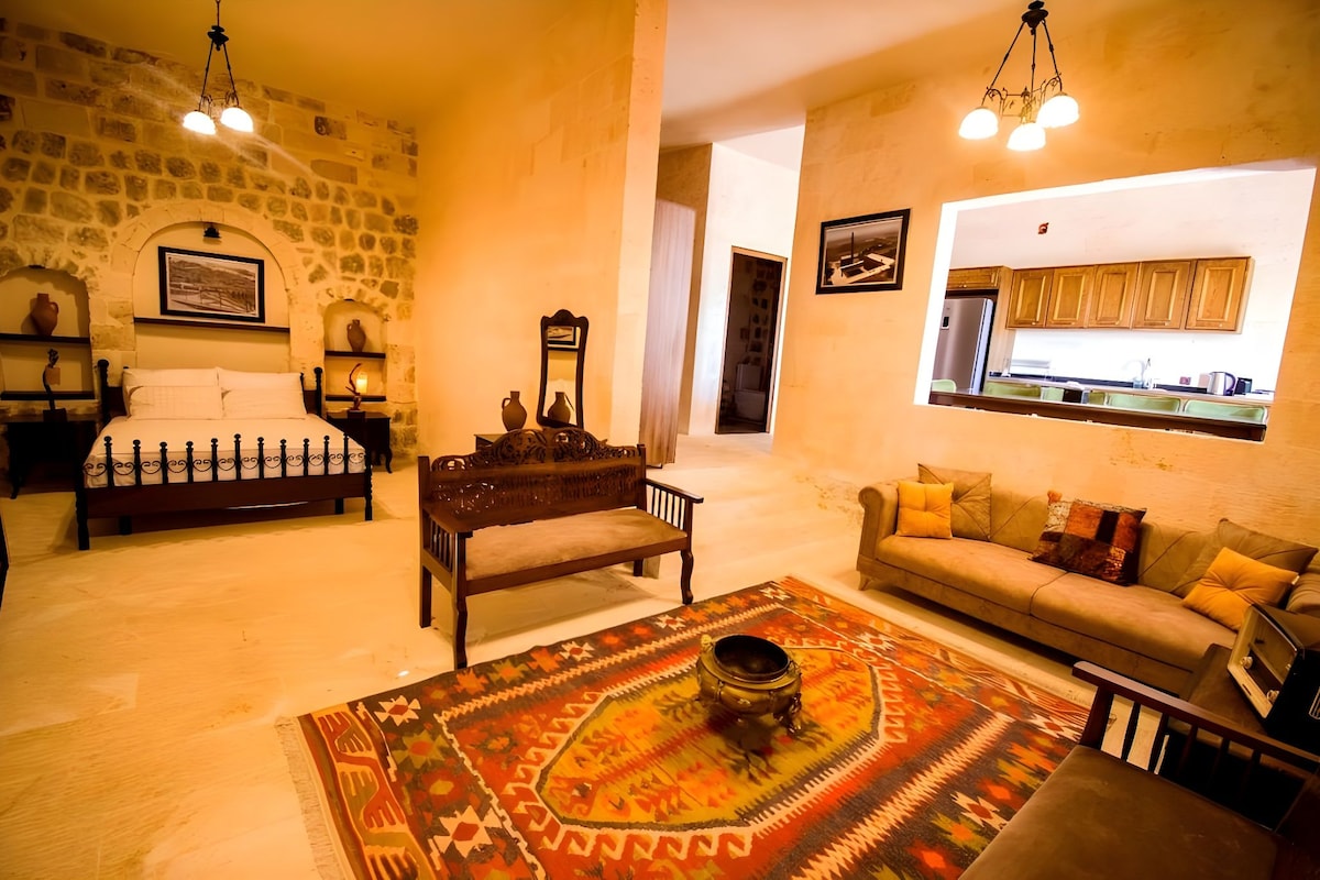 Una sala de estar acogedora con una pared de piedra, una cama, un sofá, un tapete estampado y una cocina visible a través de un arco.