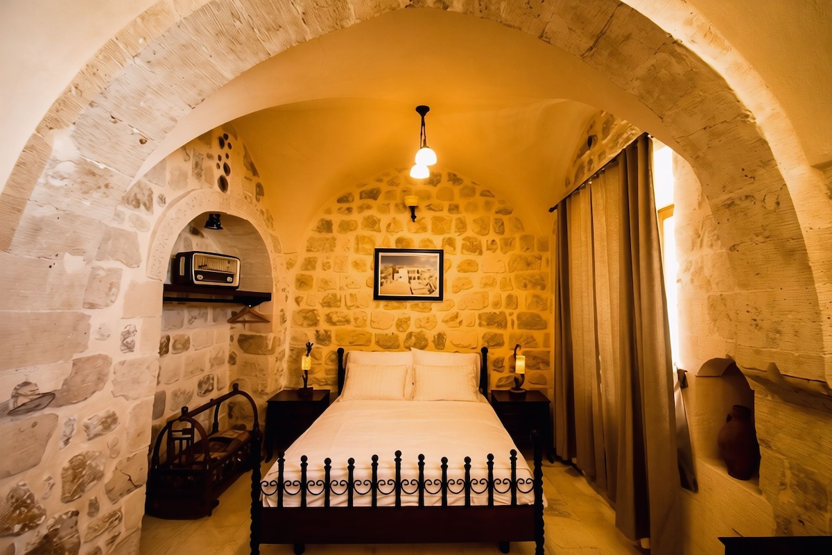 Una habitación con pared de piedra, una cama con cabecera, dos mesitas de noche, una lámpara colgante y un cuadro en la pared.