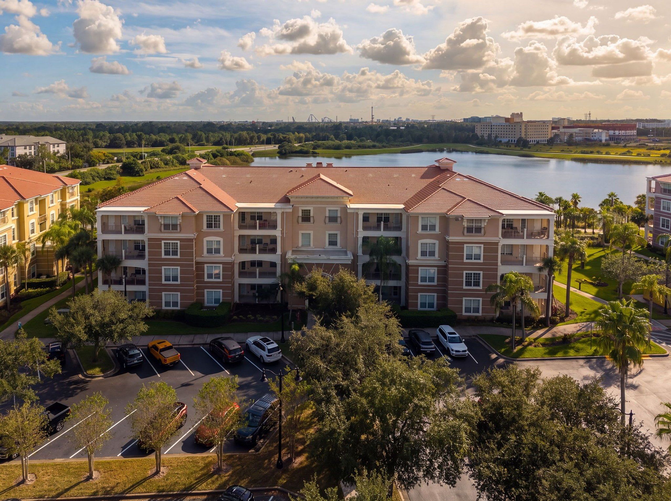 A multi-story residential building with a red-tiled roof, surrounded by a parking lot and palm trees, with a lake and cityscape in the background.