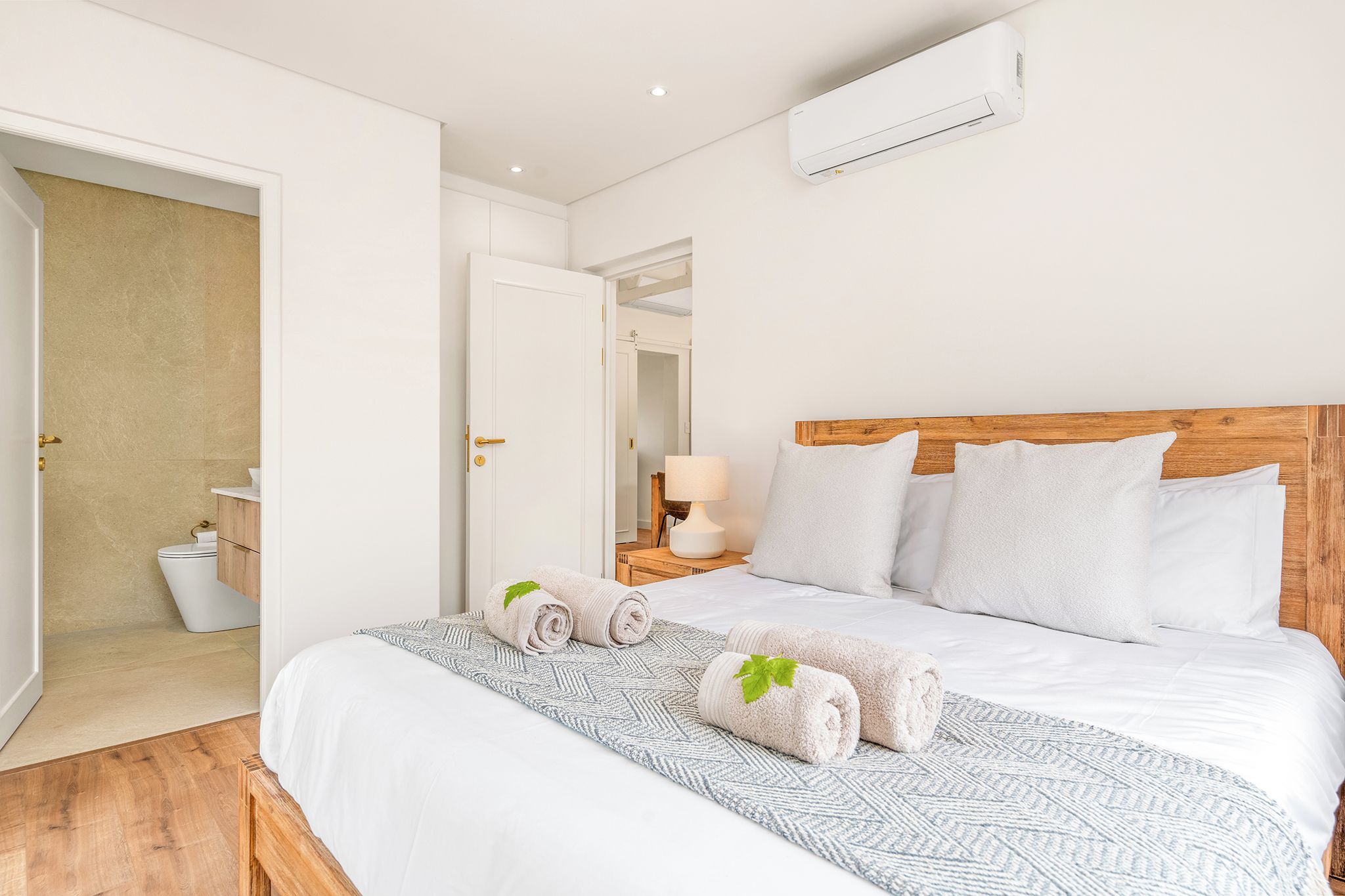 A hotel room with a bed, a bedside table, a lamp, and an air conditioning unit.
