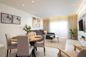 A modern living room with a dining area, grey sofa, and a flat-screen TV mounted on the wall. - Fado Apartment | Pool, Balcony & Near the Beach – Albufeira (Albufeira)