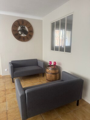 Una sala de estar con un sofá gris, un barril de madera con dos copas de vino tinto, y un reloj de pared.