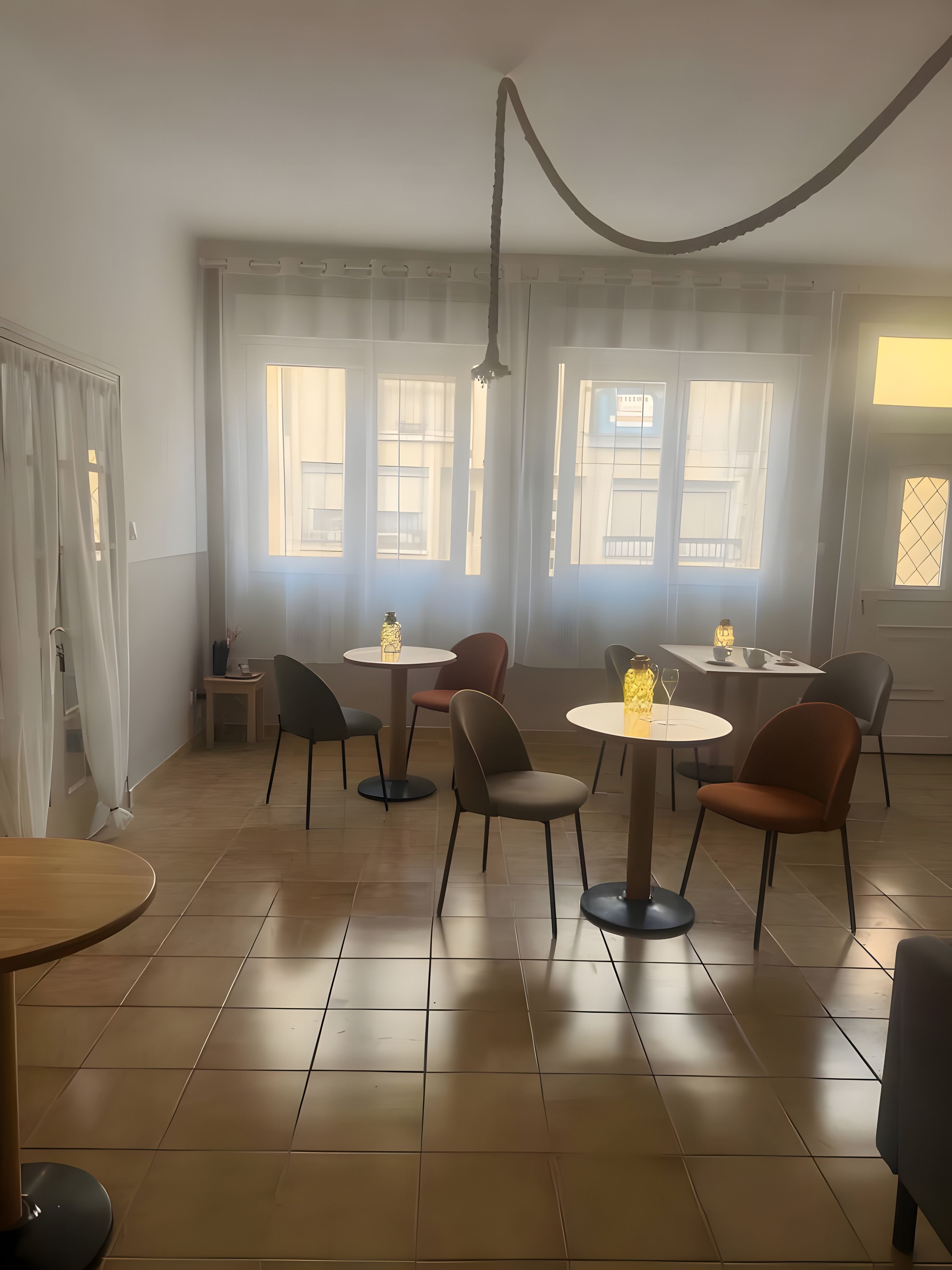 A dining area with round tables and chairs, large windows with curtains, and a tiled floor.