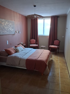 Un dormitorio con una cama, dos sillas, una mesita, una lámpara y una ventana con cortinas.