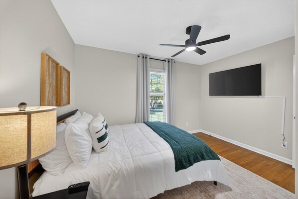 A bedroom with a bed, a ceiling fan, a TV mounted on the wall, and a window with curtains. - Bluegrass Springdale | Air Hockey & Darts (Owensboro)