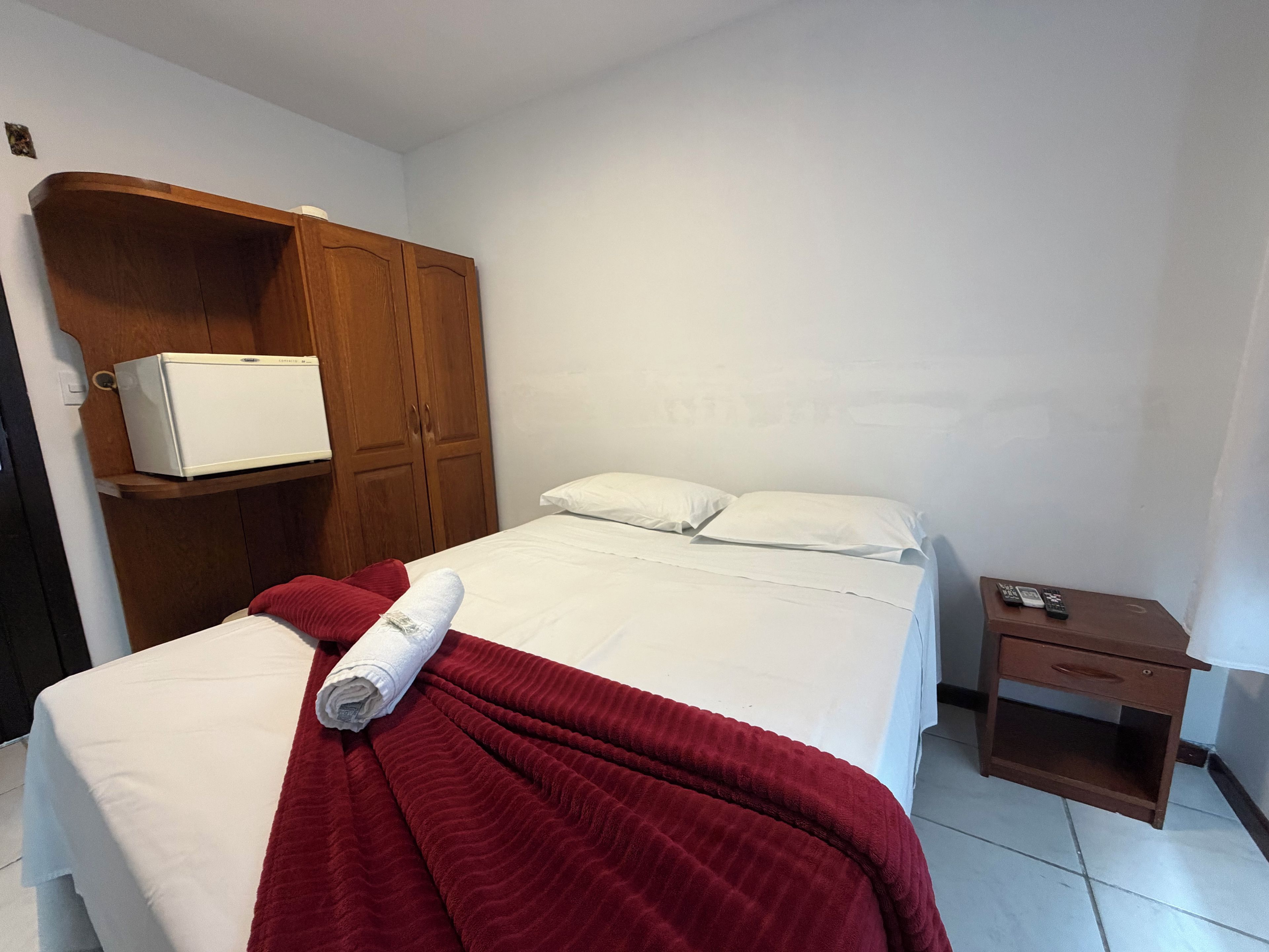A hotel room with a bed, a wooden wardrobe, a small bedside table, and a white air conditioning unit.