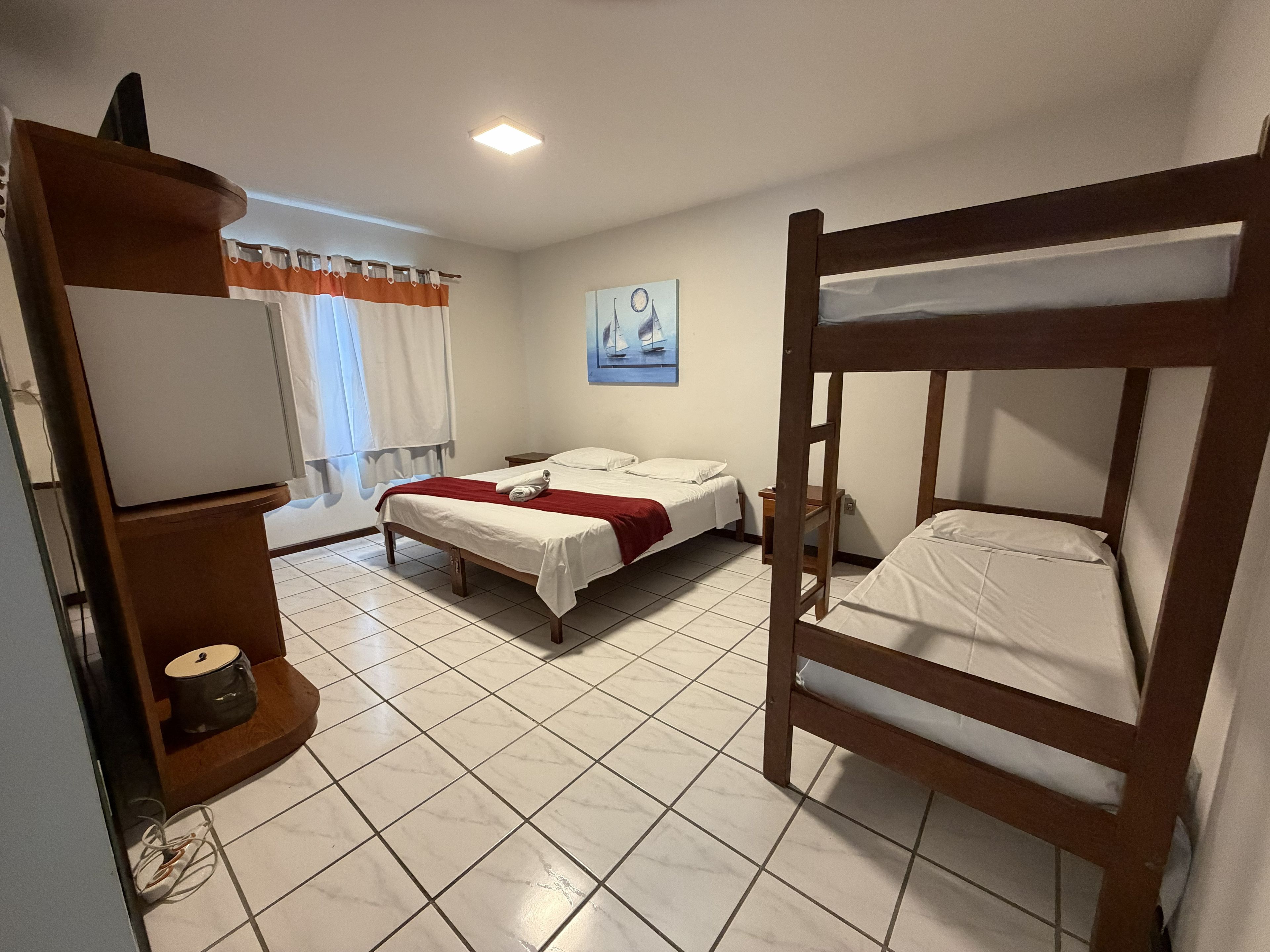 Quarto de hotel com beliche, uma cama de solteiro, escrivaninha com luminária, janela com cortinas e um quadro na parede.