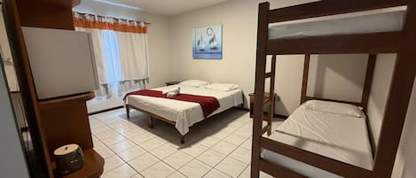Habitación de hotel con literas, una cama individual, un escritorio con lámpara, una ventana con cortinas y un cuadro en la pared.