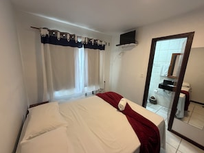 Ett hotellrum med en säng, en tv som är monterad på väggen, ett badrum som syns genom en öppen dörr och ett fönster med gardiner.