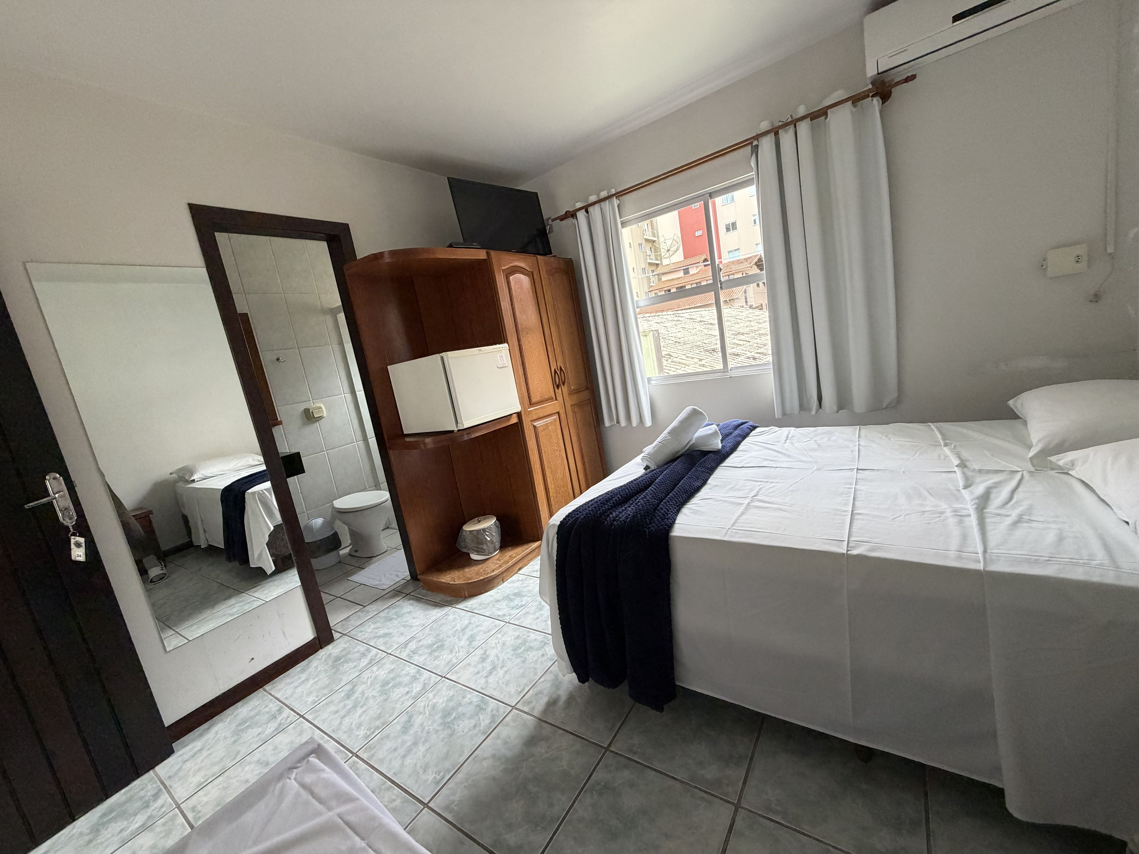 A hotel room with a bed, a wardrobe, a mirror, a toilet, and a window.