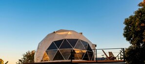 A geodesic dome structure with a transparent roof, surrounded by a metal railing, and a chair visible in the foreground. - Modern Dome Stay Near the Beach with Mountain views (San Alfonso)