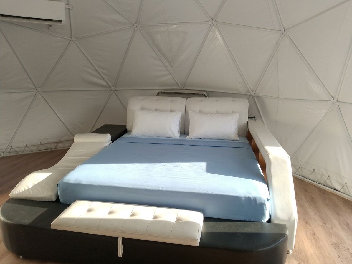 Een bed met een blauwe matras, witte kussens en een bijpassend pouf in een tentachtige kamer met een geometrisch plafondpatroon.