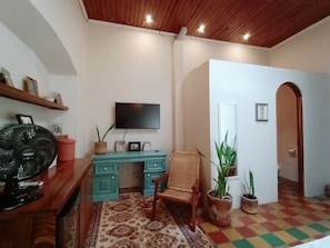 Een kamer met een houten plafond, een aan de muur bevestigde televisie, een bureau met stoel, een ventilator en diverse potplanten.