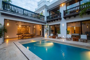Piscina con sillones de descanso y zona de bar, rodeada por un edificio de dos plantas con balcones.