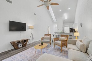 A modern living room with a flat-screen TV, wooden furniture, a ceiling fan, and a kitchen in the background. - Cozy Gainesville Apt • 2BR 2BA Stay near UF Campus (Gainesville)