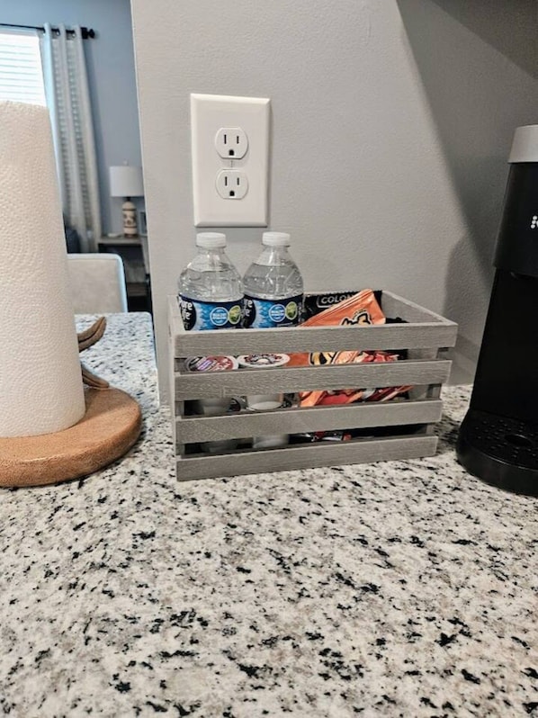 Una encimera de cocina con un caja gris que contiene botellas de agua y paquetes de snacks, un enchufe y una cafetera.