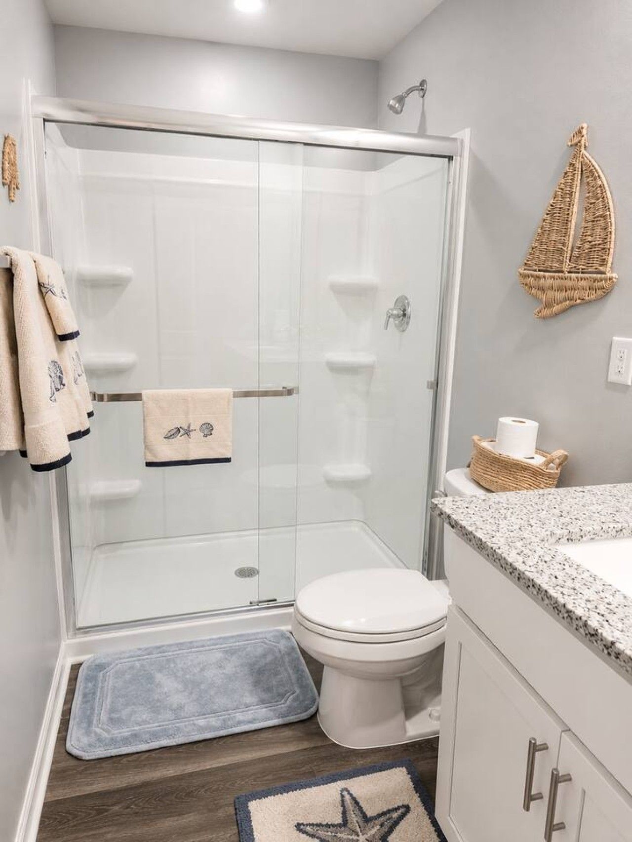 Une salle de bain moderne avec une douche à parois de verre, des toilettes blanches, un lavabo doté d’un plan de travail en granit et un tapis orné d’un motif en forme d’étoile de mer.