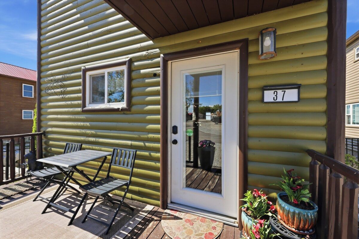 Une cabane en rondins portant le numéro 37, avec une porte, une fenêtre et une terrasse équipée d’une table et de chaises.