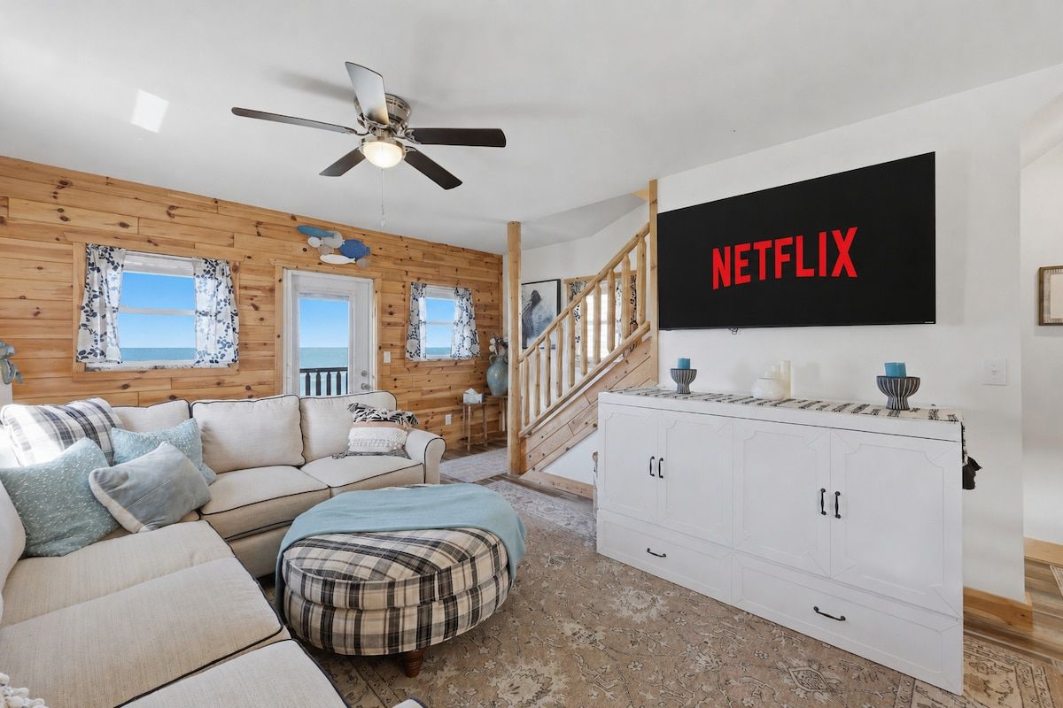 A cozy living room with a sofa, ottoman, and a TV mounted on the wall displaying 'NETFLIX'.
