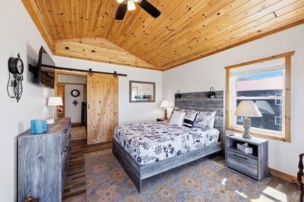 Een slaapkamer met een bed, nachtkastjes, een houten plafond, een raam en een deur naar een andere kamer.