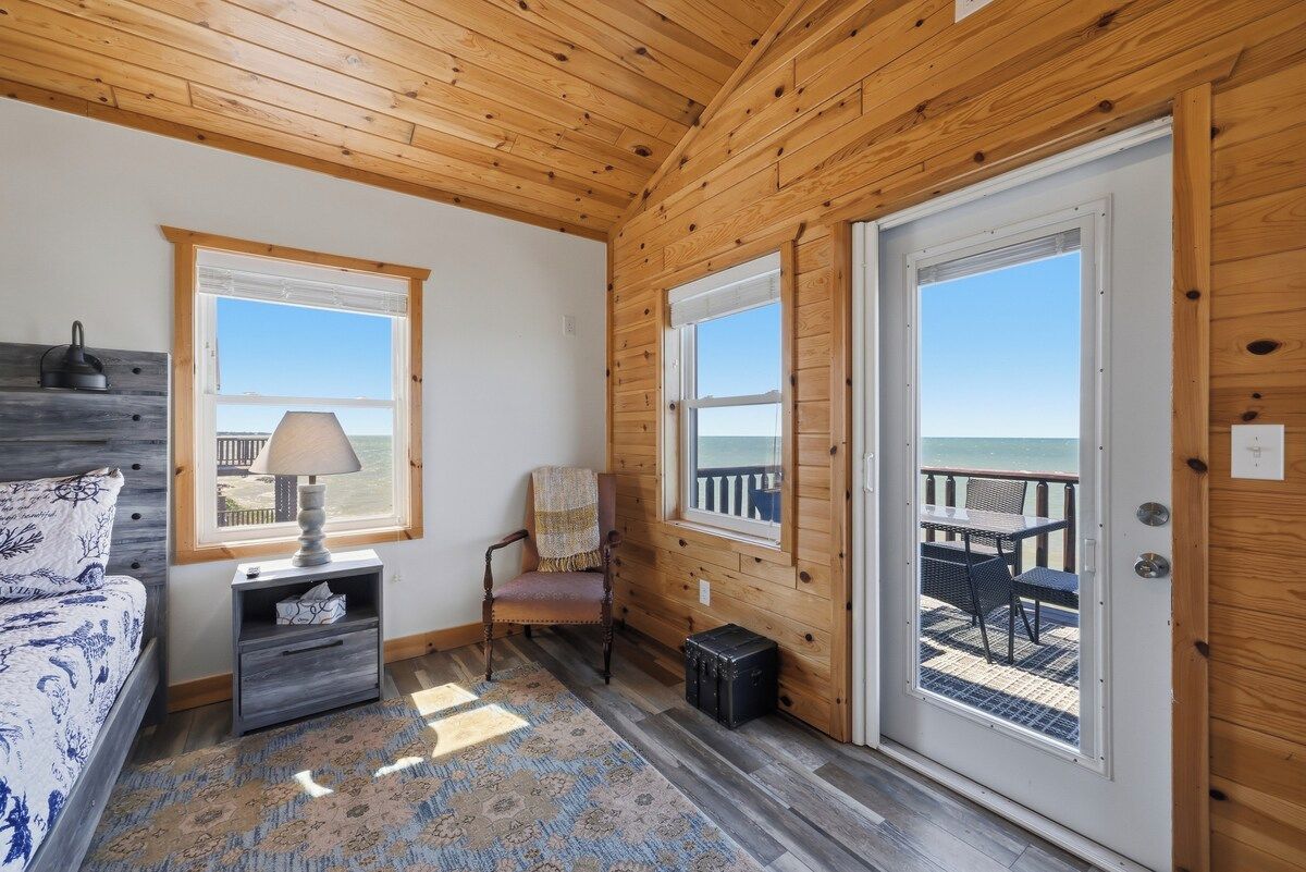Une pièce aux murs en bois, un lit recouvert d’une couvre-lit à motifs, une table de chevet avec une lampe, une chaise et une vue sur la plage par la porte.