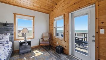 Une pièce aux murs en bois, un lit recouvert d’une couvre-lit à motifs, une table de chevet avec une lampe, une chaise et une vue sur la plage par la porte.