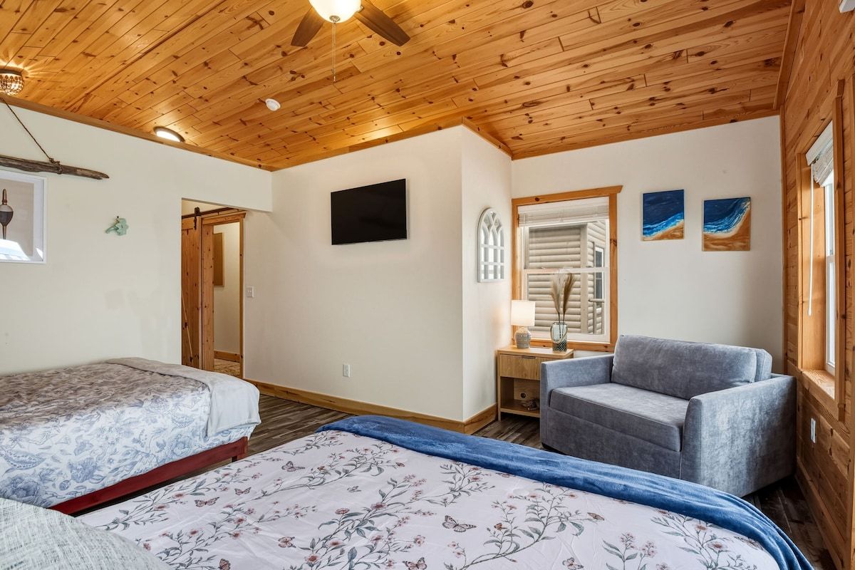 A room with wooden ceiling, a bed with floral bedding, a grey sofa, a TV, and a window with shutters.