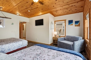 A room with wooden ceiling, a bed with floral bedding, a grey sofa, a TV, and a window with shutters. - Triple Tides: 3 Hot Tubs/Sleeps 30+/ Waterfront (Ashtabula)