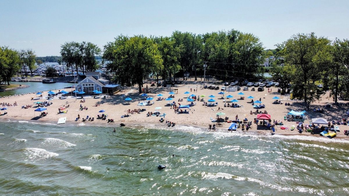 Une plage avec de nombreux parasols et des gens, un bateau amarré près du rivage, et un bâtiment avec un toit bleu.