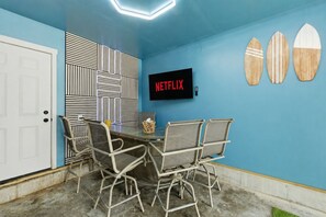 A room with a blue wall, a white door, a table with chairs, a TV screen displaying 'NETFLIX', and three surfboard decorations.