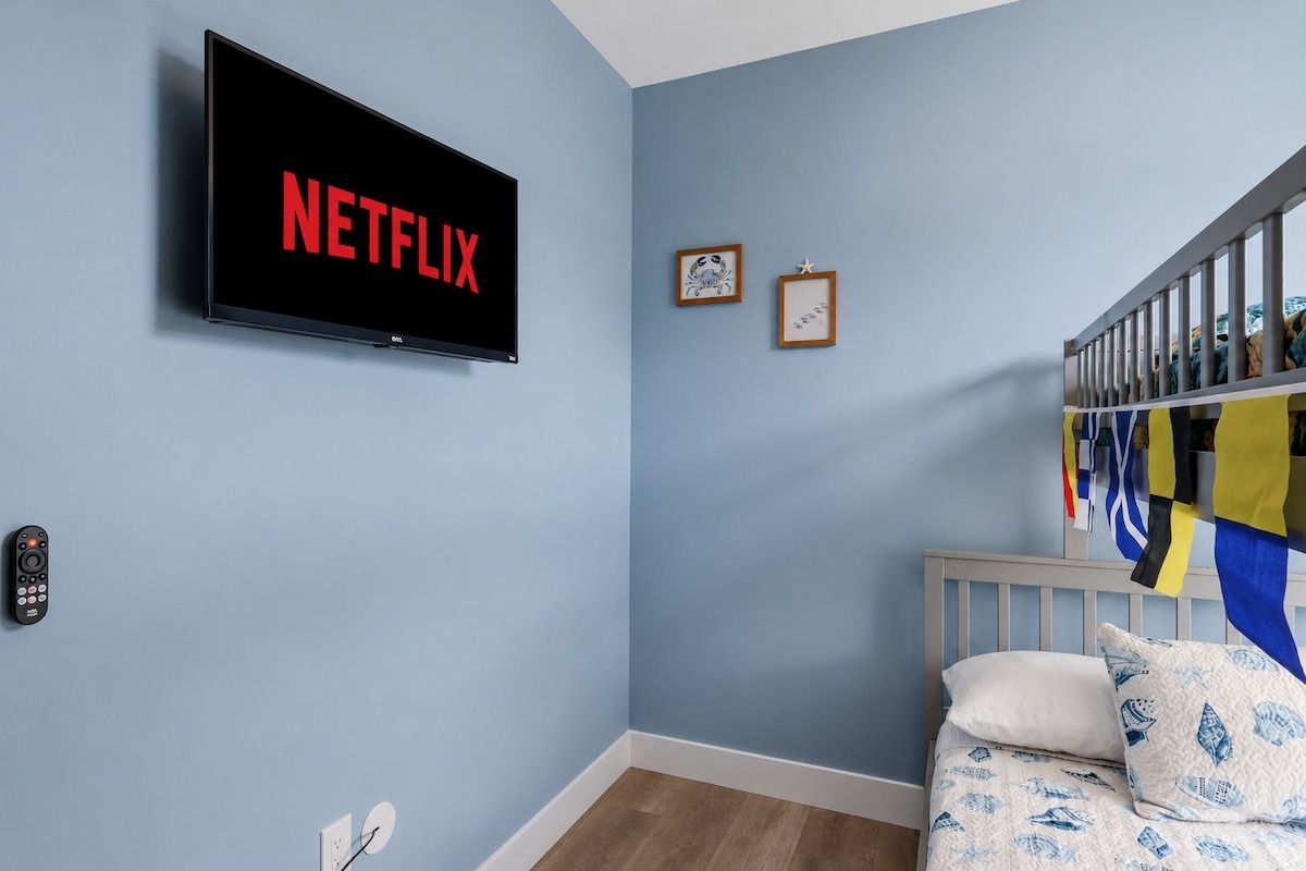 A room with a bed, a wall-mounted TV displaying 'NETFLIX', a framed picture, and a flag.