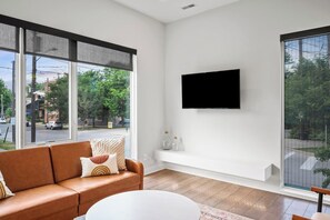 A modern living room with a large window, a flat-screen TV mounted on the wall, a brown leather sofa, and a white coffee table. - Twin Terraces-Skyline Views with Two Rooftop Decks (Cleveland)