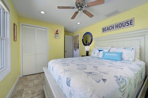 Ein Schlafzimmer mit einem Bett, einem Deckenventilator, einem Wandzeichen mit der Aufschrift 'BEACH HOUSE' und einer Tür mit Klappläden.