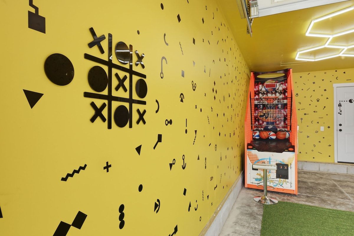 A room with a yellow wall decorated with black geometric shapes and symbols, a small table with a chair, and a basketball arcade game.