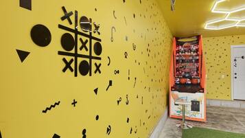 A room with a yellow wall decorated with black geometric shapes and symbols, a small table with a chair, and a basketball arcade game.