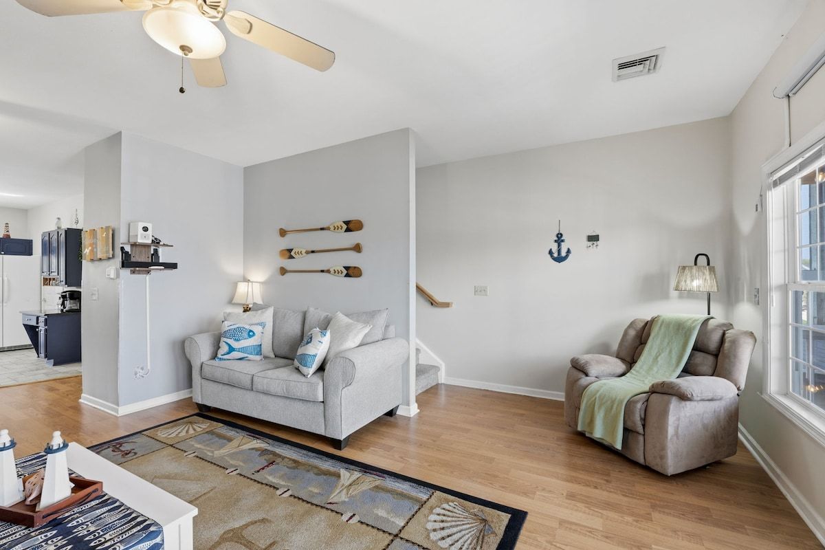 A living room with a grey sofa, a massage chair, a patterned rug, and nautical decor including wall-mounted anchors and a ship's wheel.