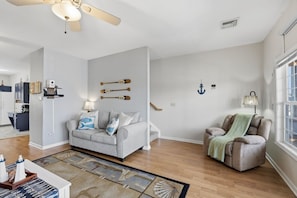Een woonkamer met een grijze bank, een massagestoel, een getextureerd vloerkleed en nautische decoratie, waaronder aan de muur bevestigde ankers en een scheepsschijf.