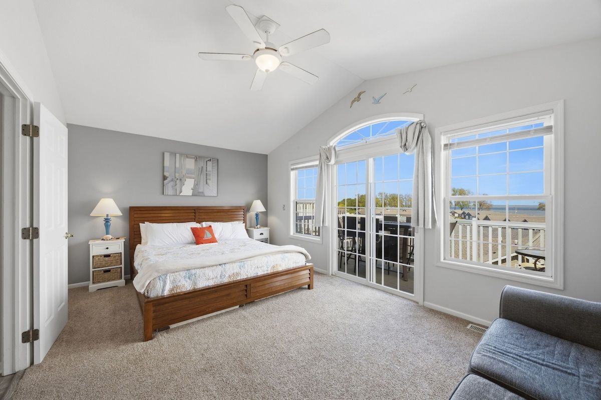 A bedroom with a bed, a ceiling fan, a nightstand, a lamp, and a view through a large window.