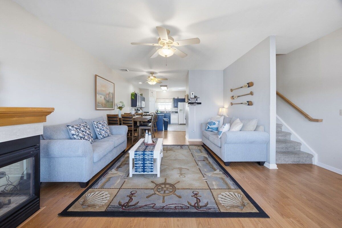 A spacious living room with a fireplace, two sofas, a coffee table, and a nautical-themed rug. A kitchen and dining area are visible in the background.