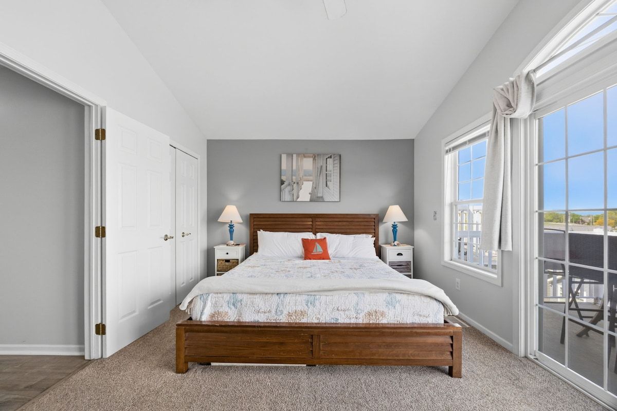 A bedroom with a bed, two bedside tables, a nightstand, a window with curtains, and a view of the outside.
