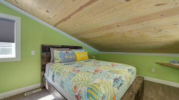 Ein Schlafzimmer mit schrägen Wänden, einem Bett mit farbenfroher Bettdecke, einem Kopfteil und einem Fenster mit Jalousien.