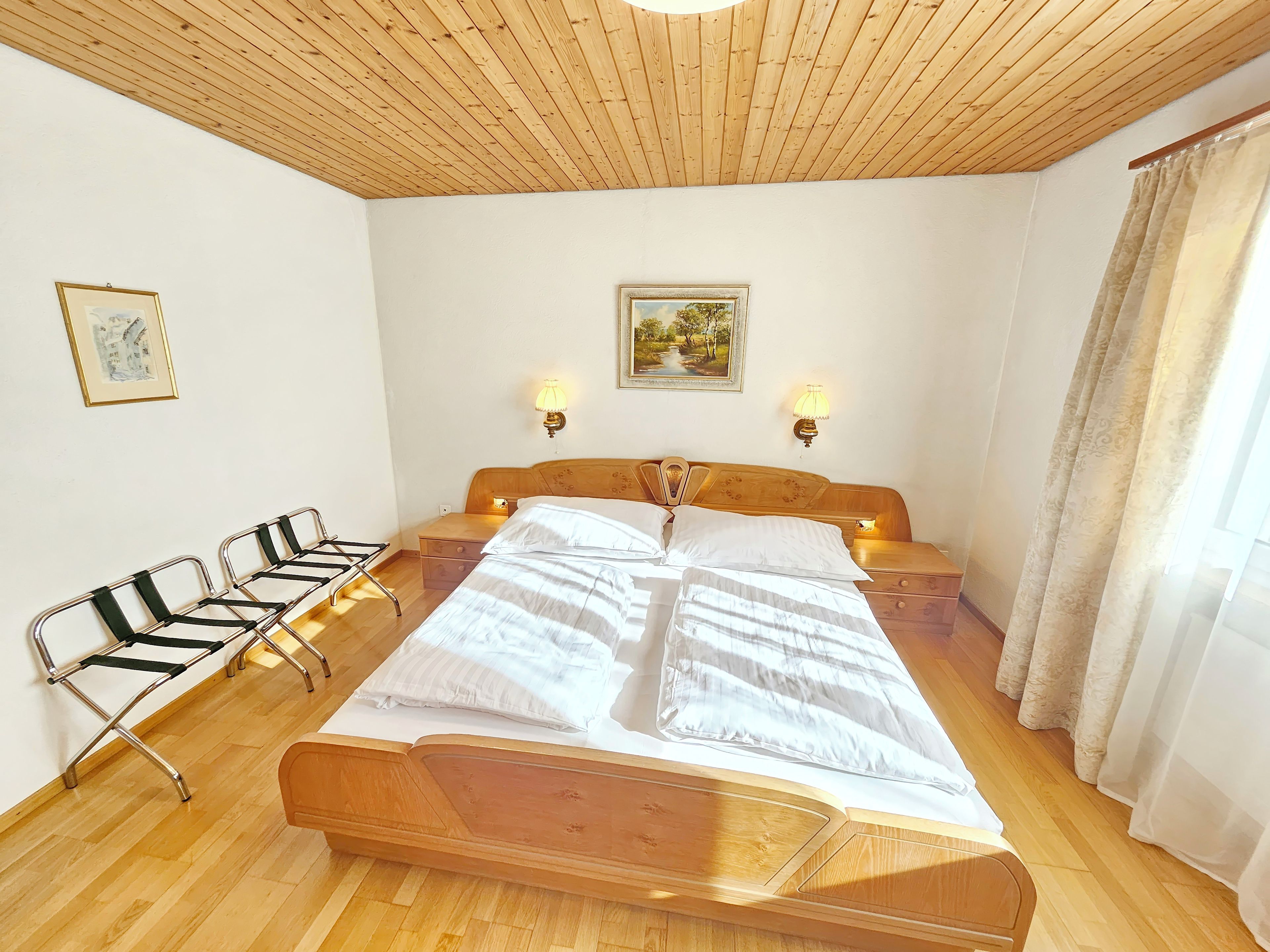 Ein Schlafzimmer mit Holzbalkendecke, einem großen Bett, zwei Nachttischen, einem Beistelltisch, einem Sessel und zwei Wandleuchten.