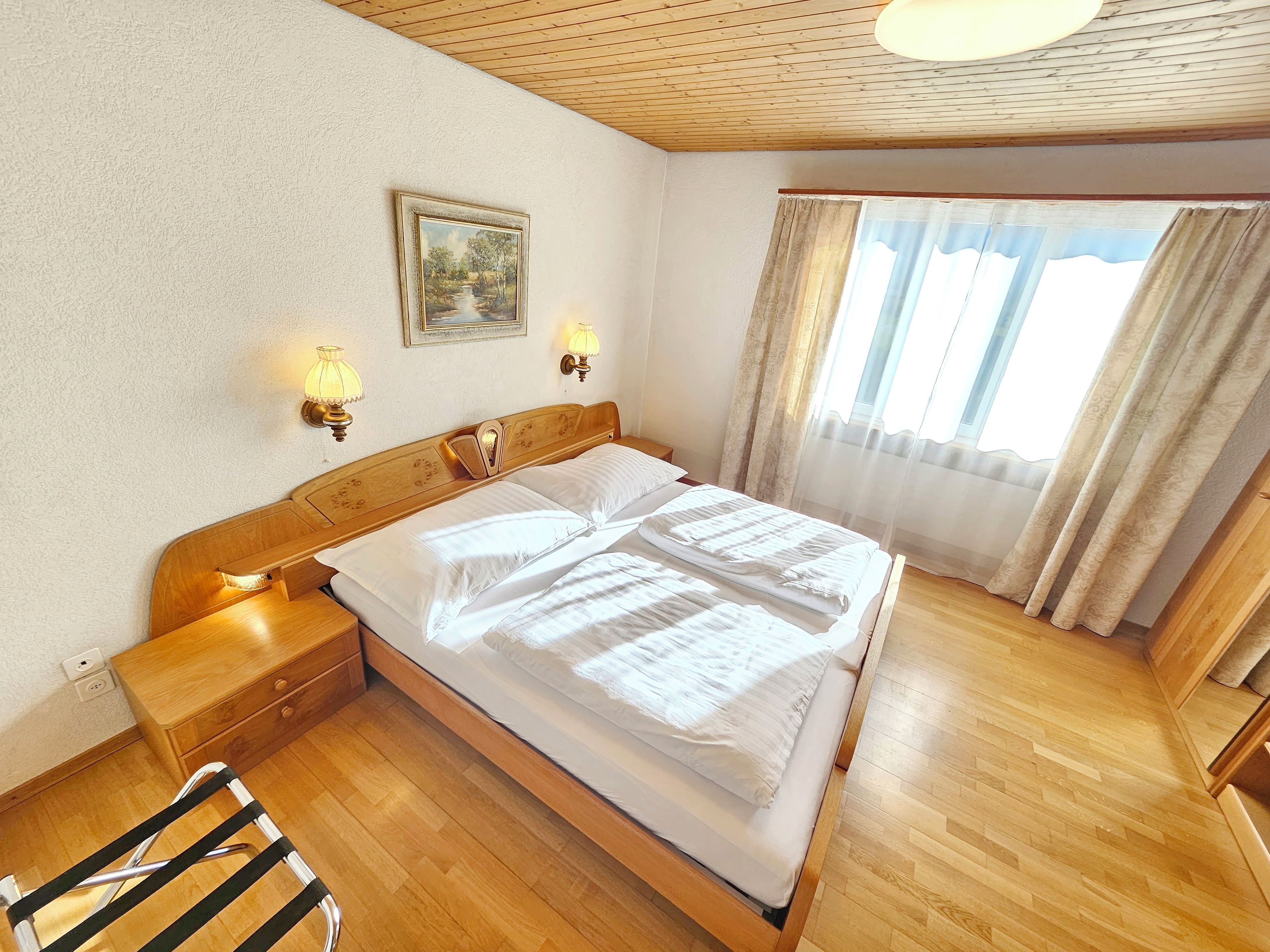 Ein Hotelzimmer mit einem hölzernen Bett, Nachttischlampen, einem Gemälde an der Wand und einem Fenster mit Vorhängen.