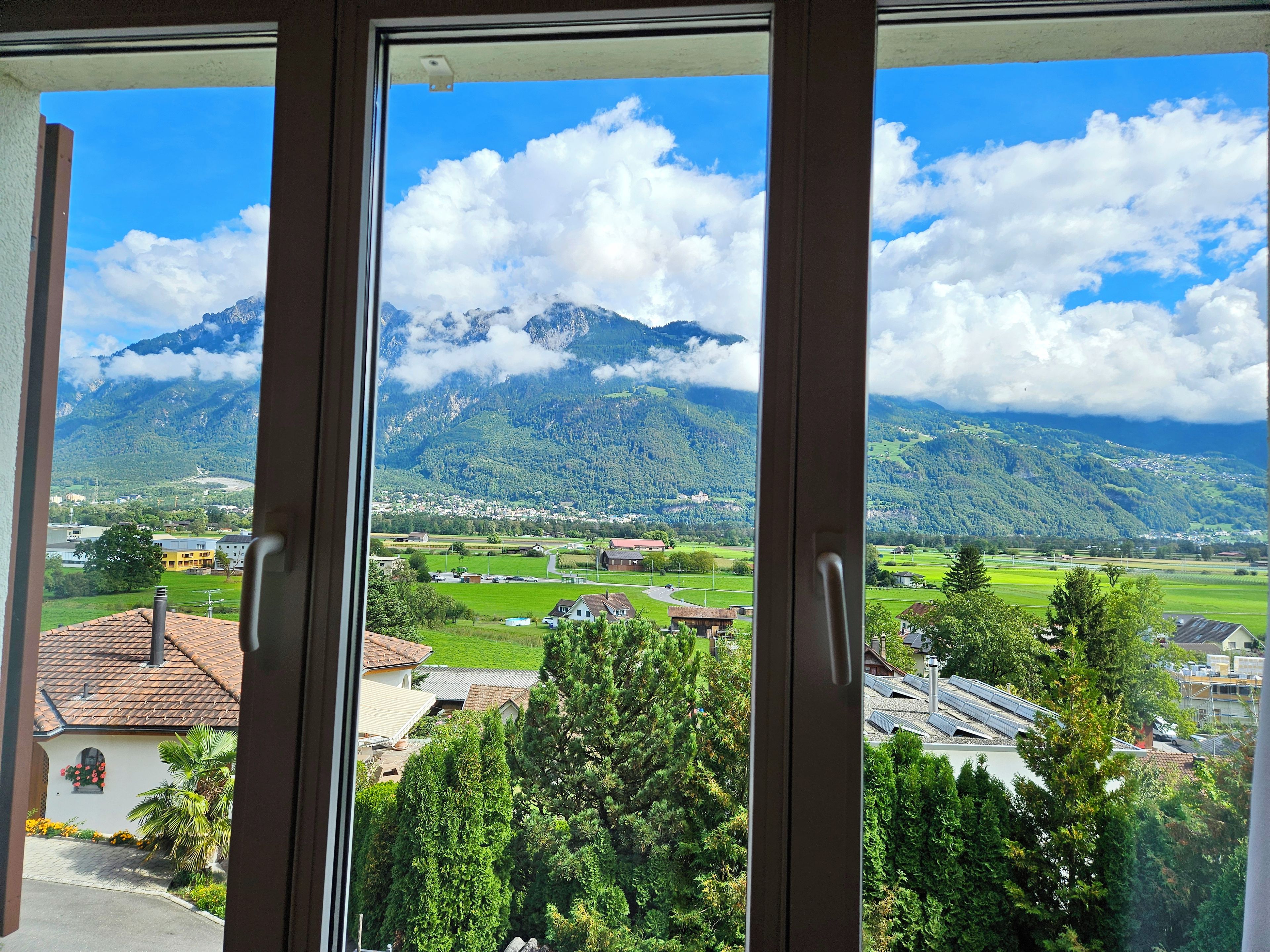 Vista a través de una ventana que muestra un paisaje montañoso, incluyendo un pico destacado y un valle con edificaciones.