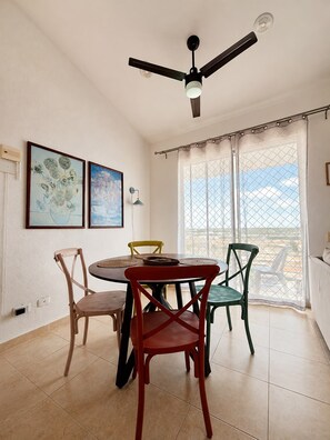Un coin repas avec une table ronde, quatre chaises, un ventilateur de plafond, deux tableaux accrochés au mur et une grande fenêtre donnant sur un beau paysage.