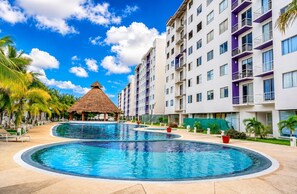 Un complexe hôtelier doté d’une piscine centrale, d’une structure à toit de chaume et d’immeubles modernes à plusieurs étages, chacun doté de balcons.