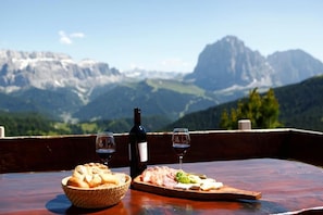 Uma mesa com uma garrafa de vinho, duas taças, uma cesta de pão e uma tábua de madeira com comida, com montanhas ao fundo.
