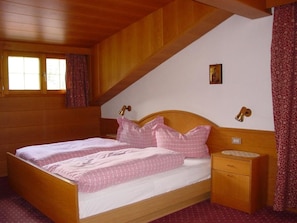 A room with a wooden bed, a nightstand, a window with curtains, and a small picture on the wall. - Garni Conturina - B&B (Santa Cristina Valgardena)